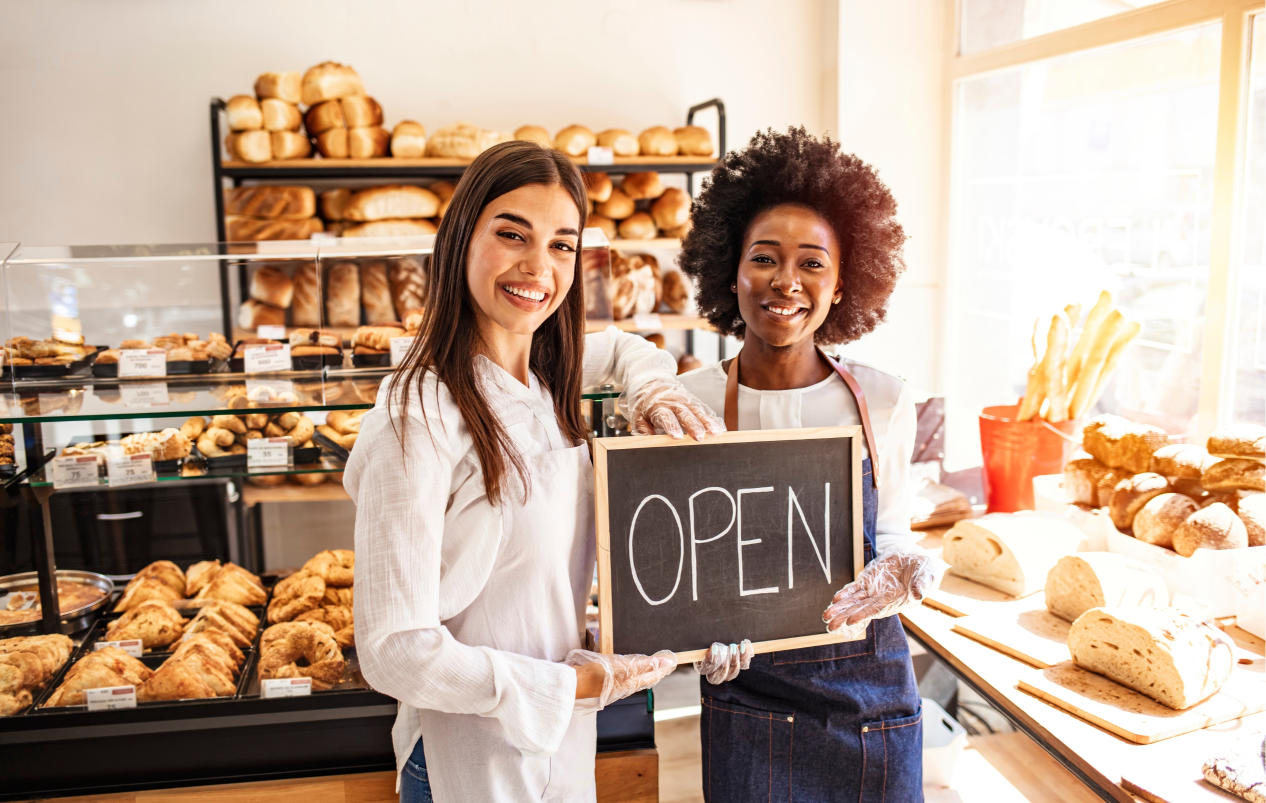 A White Woman and a Black Woman in a Bakery Holding an Open Sign